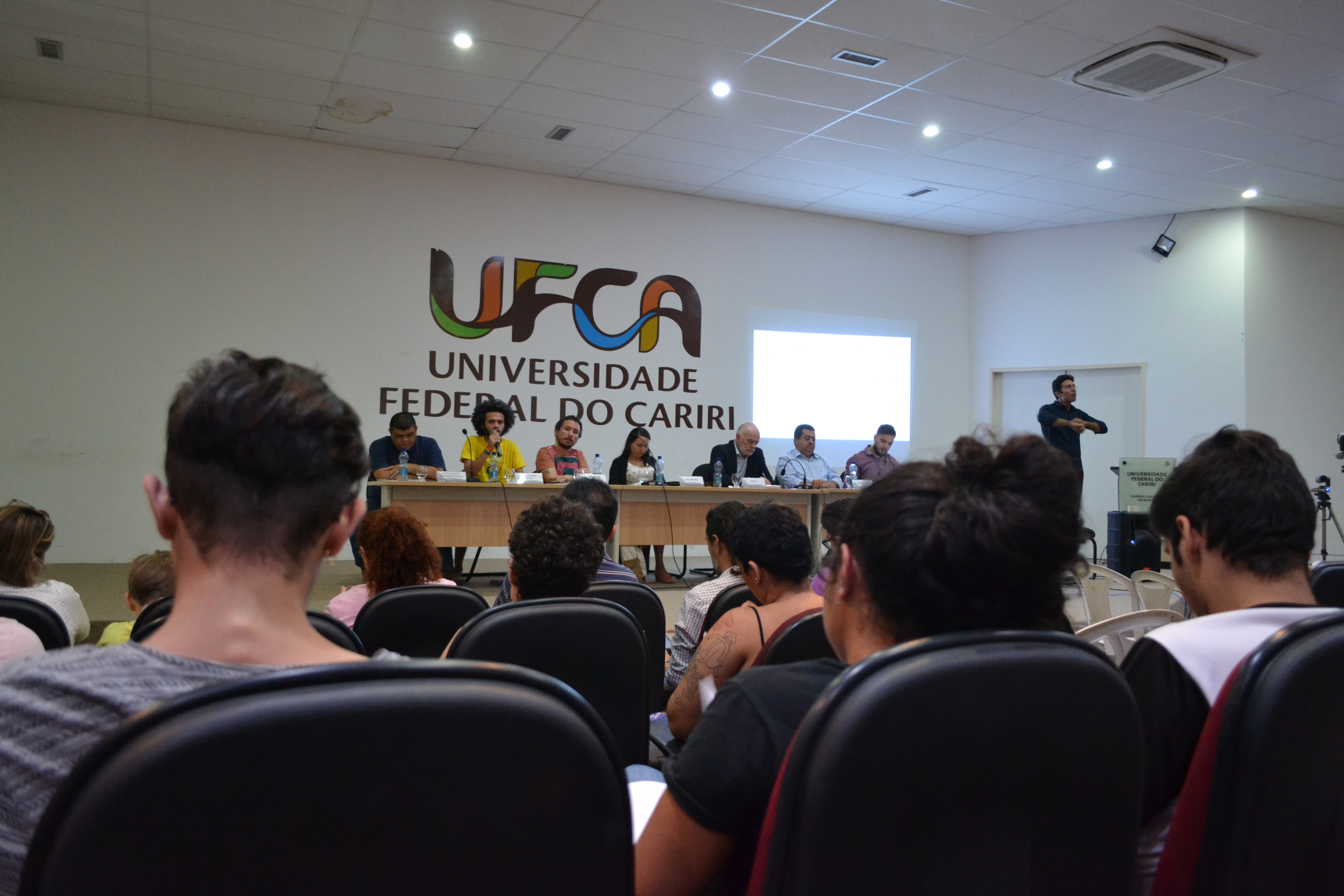 Em um auditório, pessoas estão assistindo a discussão da assembleia. No palco, mesa formada por representantes da comunidade acadêmica.